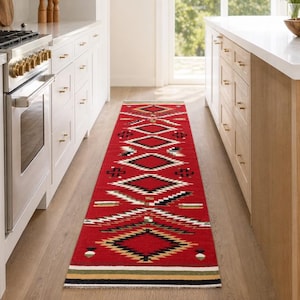 May include: A long, rectangular runner rug featuring a vibrant red base color with a repeating geometric pattern in black, white, and tan. The design includes diamond shapes and horizontal stripes, finished with a simple border at the ends.
