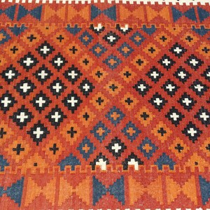 May include: A woven rug featuring a repeating geometric pattern of diamonds and crosses in shades of burnt orange, deep blue, black, and white. The design consists of two large central diamond motifs filled with smaller cross shapes.