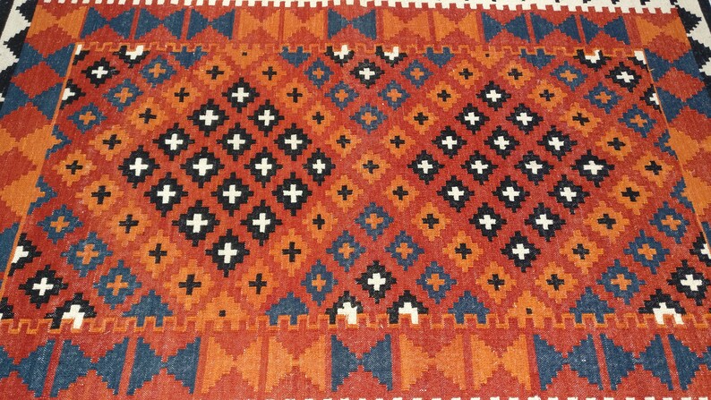 May include: A woven rug featuring a repeating geometric pattern of diamonds and crosses in shades of burnt orange, deep blue, black, and white. The design consists of two large central diamond motifs filled with smaller cross shapes.