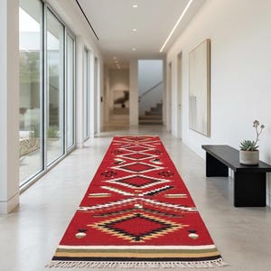 May include: A long, rectangular runner rug featuring a vibrant red base with a repeating geometric pattern in black, white, and tan. The design includes diamond shapes and horizontal stripes, finished with a short, light-colored fringe at both ends.