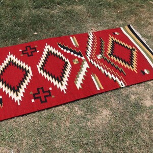 May include: A rectangular woven rug featuring a vibrant red background with a repeating geometric pattern of diamonds and crosses in black, white, and olive green. The rug has a fringed edge with horizontal stripes in black, white, and olive green.