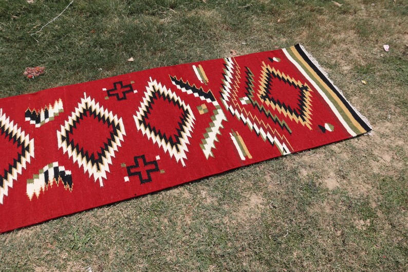 May include: A rectangular woven rug featuring a vibrant red background with a repeating geometric pattern of diamonds and crosses in black, white, and olive green. The rug has a fringed edge with horizontal stripes in black, white, and olive green.