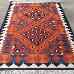 May include: A rectangular area rug featuring a geometric pattern in shades of orange, blue, black, and white. The central field displays a repeating diamond motif with small white crosses, surrounded by a border of triangles and fringe on both ends.