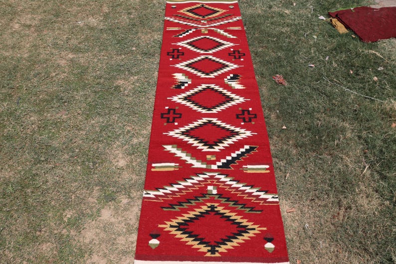 May include: A long, rectangular runner rug featuring a vibrant red background with a repeating geometric pattern. The design includes stacked diamonds, cross shapes, and linear accents in contrasting colors of black, white, and tan throughout.