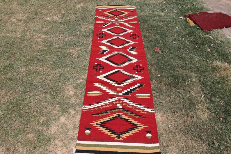 May include: A long, rectangular runner rug featuring a vibrant red base color. The rug is decorated with a repeating geometric pattern consisting of diamonds, crosses, and jagged lines in black, white, tan, and green, finished with striped borders.