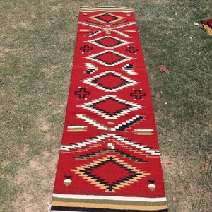 May include: A long, rectangular runner rug featuring a vibrant red base color. The rug is decorated with a repeating geometric pattern consisting of diamonds, crosses, and jagged lines in black, white, tan, and green, finished with striped borders.
