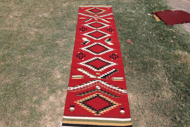 May include: A long, rectangular runner rug featuring a vibrant red base color. The rug is decorated with a repeating geometric pattern consisting of diamonds, crosses, and jagged lines in black, white, tan, and green, finished with striped borders.