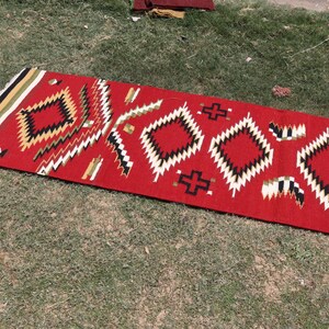 May include: A rectangular runner rug featuring a vibrant red base color with a repeating geometric pattern of diamonds and crosses in black, white, and tan. The rug has a fringed edge with horizontal stripes in black, white, and tan colors.