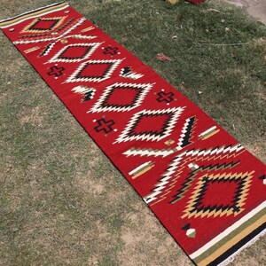May include: A long, rectangular runner rug featuring a vibrant red base color with a repeating geometric pattern of diamonds and crosses in black, white, and tan. The rug has a flat-woven texture and includes fringed ends with striped borders.