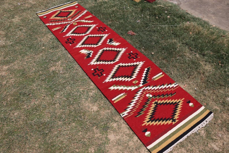 May include: A long, rectangular runner rug featuring a vibrant red base color with a repeating geometric pattern of diamonds and crosses in black, white, and tan. The rug has a flat-woven texture and includes fringed ends with striped borders.