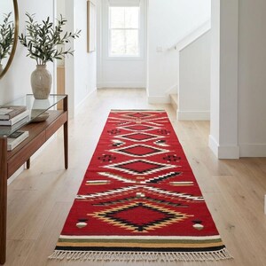May include: A long rectangular runner rug featuring a vibrant red base with geometric patterns in black, white, and tan. The design includes repeating diamond shapes and horizontal stripes, finished with short white fringe at the ends.
