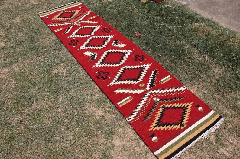 May include: A long, rectangular runner rug featuring a vibrant red base color with a repeating geometric pattern of diamonds and crosses in black, white, and tan. The rug has a flat-woven texture and includes fringed ends with striped borders.