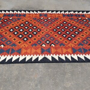 May include: A rectangular woven rug featuring a geometric pattern in shades of orange, blue, black, and white. The central field displays a repeating diamond motif with small crosses, framed by a border of triangles and finished with white fringe.