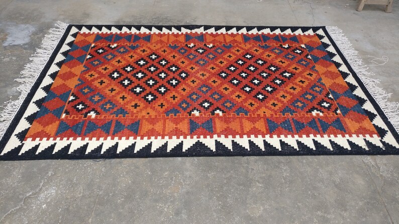 May include: A rectangular woven rug featuring a geometric pattern in shades of orange, blue, black, and white. The central field displays a repeating diamond motif with small crosses, framed by a border of triangles and finished with white fringe.