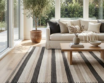 May include: A rectangular area rug featuring a pattern of vertical stripes in shades of charcoal gray, beige, and cream. The rug has a visible whip-stitched edge detail along the short ends, creating a textured border finish on the floor.
