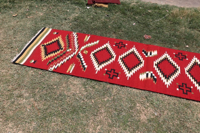 May include: A rectangular runner rug featuring a vibrant red base color with a repeating geometric pattern of diamonds and crosses in black, white, and tan. The rug has a fringed edge with horizontal stripes in black, white, and tan colors.
