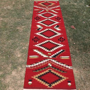 May include: A long, rectangular runner rug featuring a vibrant red background with a repeating geometric pattern. The design includes stacked diamonds, cross shapes, and linear accents in contrasting colors of black, white, and tan throughout.