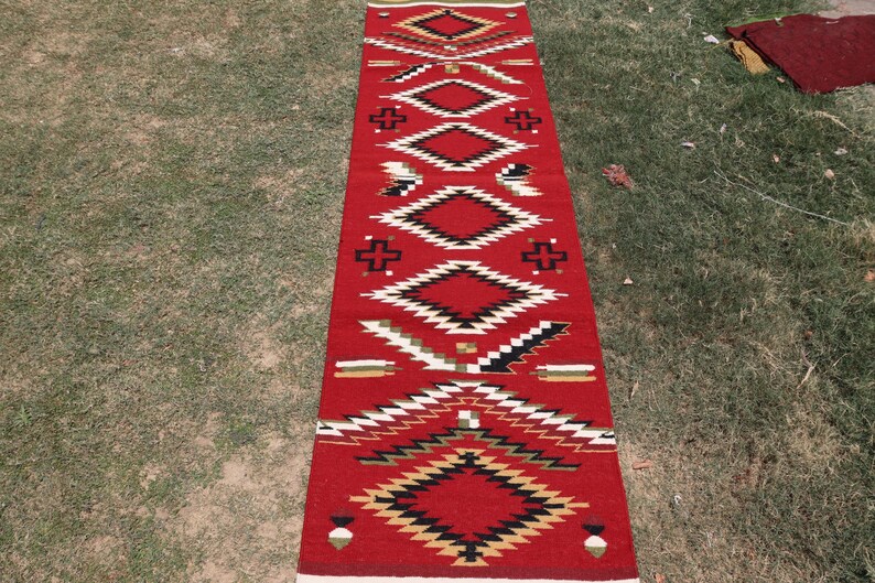 May include: A long, rectangular runner rug featuring a vibrant red background with a repeating geometric pattern. The design includes stacked diamonds, cross shapes, and linear accents in contrasting colors of black, white, and tan throughout.
