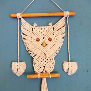 Peut inclure: Une suspension murale en macram&eacute; en forme de hibou blanche avec des chevilles en bois et un long pompon. Le hibou a deux yeux en bois.