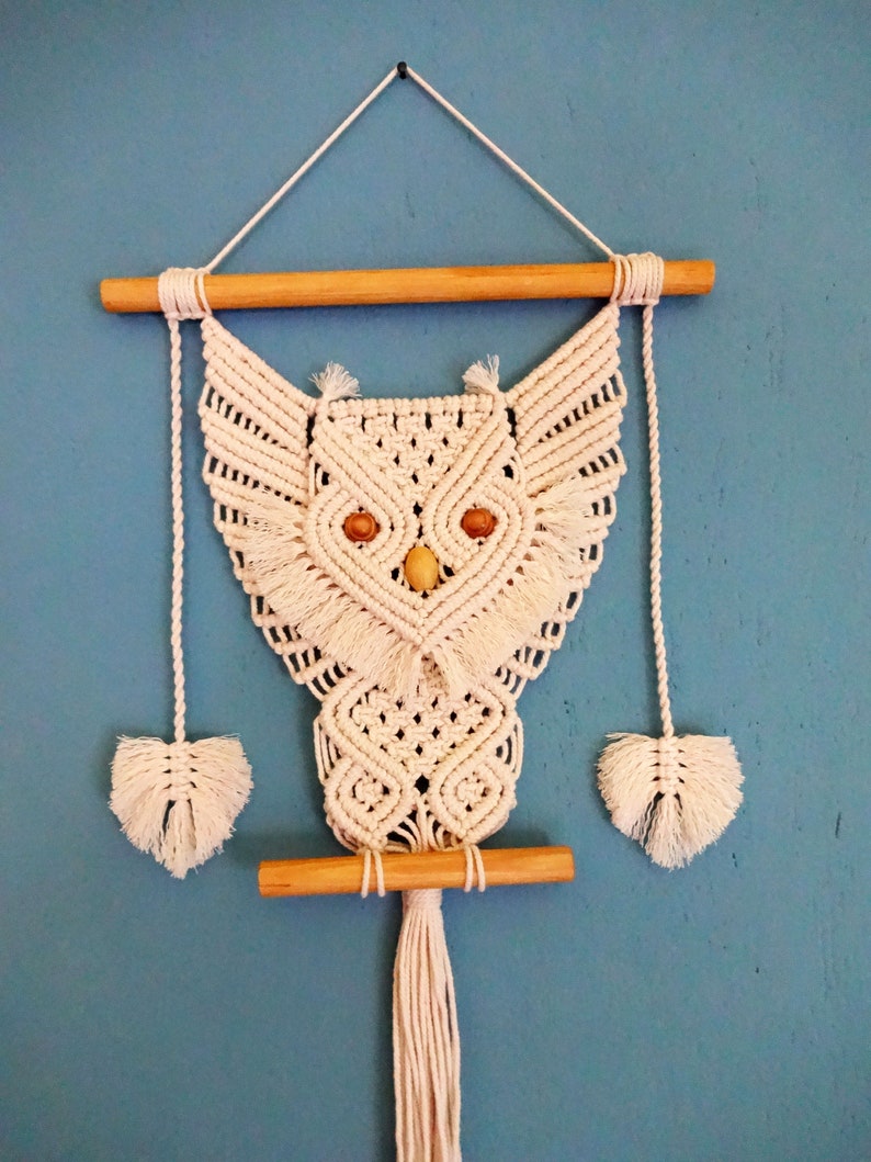 Peut inclure: Une suspension murale en macram&eacute; en forme de hibou blanche avec des chevilles en bois et un long pompon. Le hibou a deux yeux en bois.