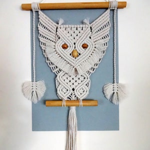 Peut inclure: Une suspension murale en macram&eacute; en forme de hibou blanche avec des chevilles en bois et une longue frange. Le hibou a des yeux en bois.