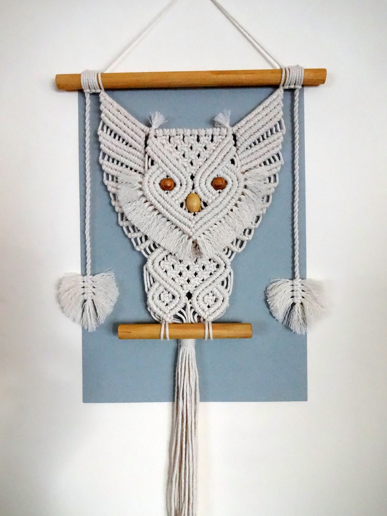 Peut inclure: Une suspension murale en macram&eacute; en forme de hibou blanche avec des chevilles en bois et une longue frange. Le hibou a des yeux en bois.