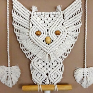 Peut inclure: Suspension murale en macram&eacute; en forme de hibou blanc avec une baguette en bois et des yeux en perles de bois. Le hibou a une frange autour de la t&ecirc;te et des ailes.
