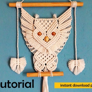 Peut inclure: Une suspension murale en macram&eacute; en forme de hibou blanche avec des chevilles en bois et des glands. Le hibou a des yeux en perles de bois. Le texte "Tutoriel" et "t&eacute;l&eacute;chargement instantan&eacute; pdf" est visible sous le hibou.