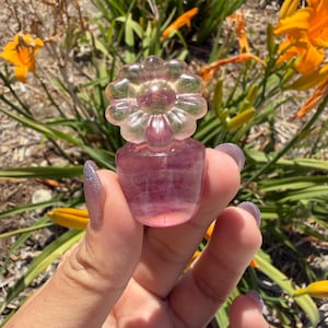 May include: A small, carved fluorite figurine shaped like a flower sitting atop a rounded base. The stone displays translucent layers of purple, pink, and pale green hues, with a smooth, polished surface that catches the sunlight.