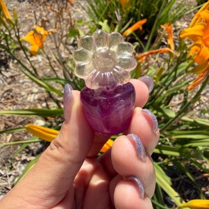 May include: A small carved stone figurine shaped like a flower with a translucent, pale yellow blossom and a deep purple, rounded base. The object is crafted from polished mineral material, showcasing natural color gradients and smooth, reflective surfaces.