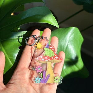 Può includere: Un portachiavi in acrilico trasparente con un design a fungo stravagante. Presenta una rana con un cappello, un sole, fiori e cristalli. Il fungo ha un cappello rosso con pois bianchi. Il portachiavi è tenuto in una mano.