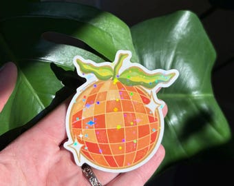 May include: A sticker featuring a disco ball design with orange and yellow squares, accented with colorful glitter. The sticker has a green stem and leaves at the top. The sticker is held in a hand in front of a large green leaf.