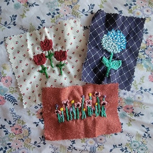 Op de afbeelding: Drie geborduurde vierkante stoffen met verschillende bloemenmotieven. Een vierkant heeft drie rode tulpen, een ander een blauwe hortensia en de laatste een rij roze bloemen.