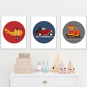 以下が含まれることがあります： 赤い飛行機、赤い車、赤い電車のイラストが描かれたカラフルな円形のフレーム付きプリントが3枚。プリントは白い壁に白いドレッサーの上に飾られています。