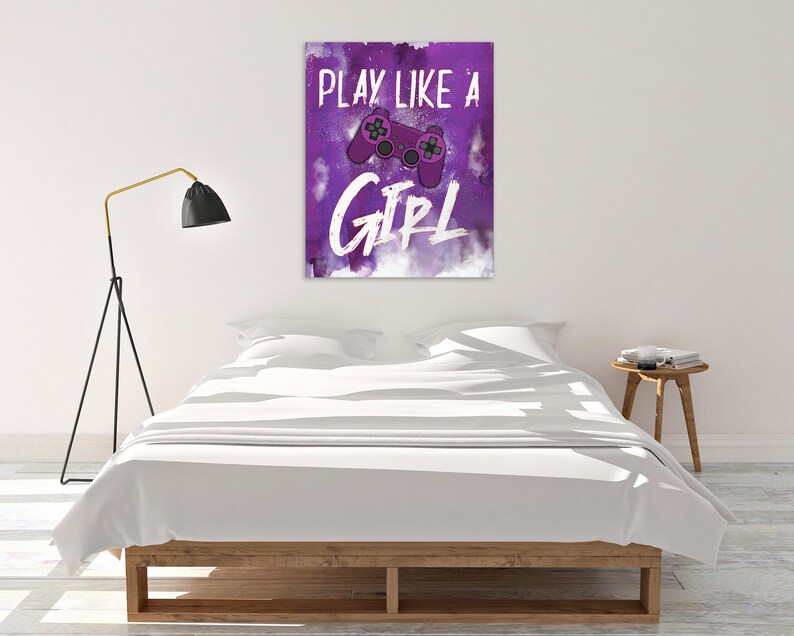 May include: Purple and white wall art with the text "PLAY LIKE A GIRL" and a purple video game controller graphic. The artwork is displayed above a bed with white bedding.