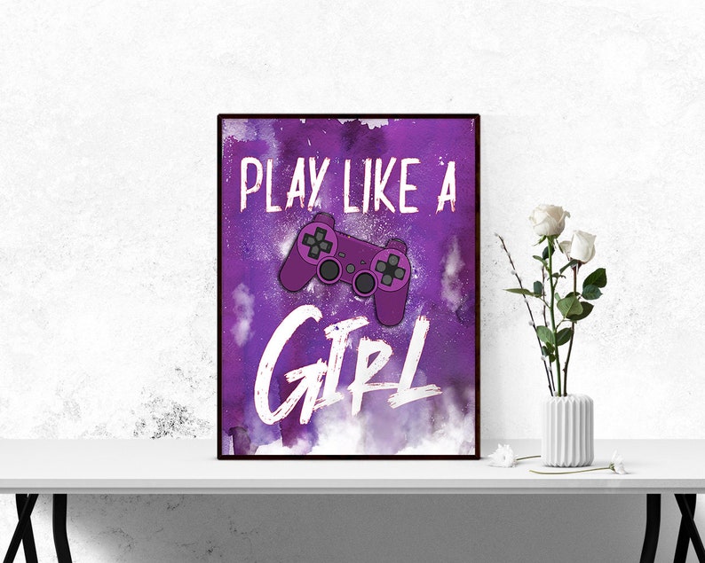 May include: Framed art print with a purple background and the words "PLAY LIKE A GIRL" in white. A purple video game controller is in the center. The print is displayed on a white surface with a vase of white roses.
