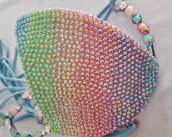 May include: A rainbow-colored face mask covered in iridescent rhinestones. The mask has light blue straps and is adorned with decorative beads. The rhinestones create a gradient effect, transitioning from green to pink.