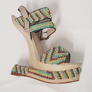 May include: A platform sandal with a wooden wedge heel. The beige straps and platform are adorned with colorful iridescent rhinestones. The heel is approximately 5 inches high.