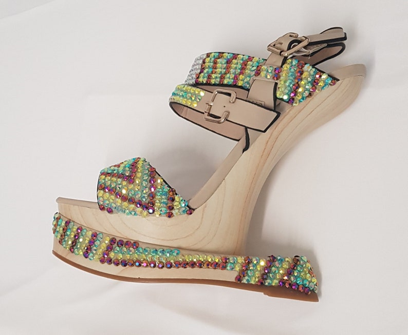 May include: A single platform sandal with a wooden wedge heel. The sandal features a beige ankle strap and toe strap, both adorned with iridescent rhinestones in a colorful pattern. The wooden platform and heel are light-colored.