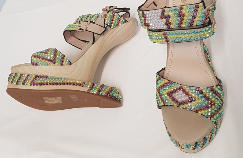 May include: A pair of platform sandals with a wooden wedge heel. The sandals feature beige straps and are embellished with colorful rhinestones in a geometric pattern. The platform and heel are made of light-colored wood.