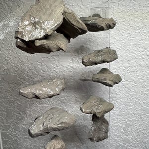May include: A unique, modern wall art installation featuring several irregularly shaped, gray-toned stone pieces. The stones are suspended at varying heights, creating a floating effect against a textured, light gray background. The arrangement suggests a natural, organic theme.