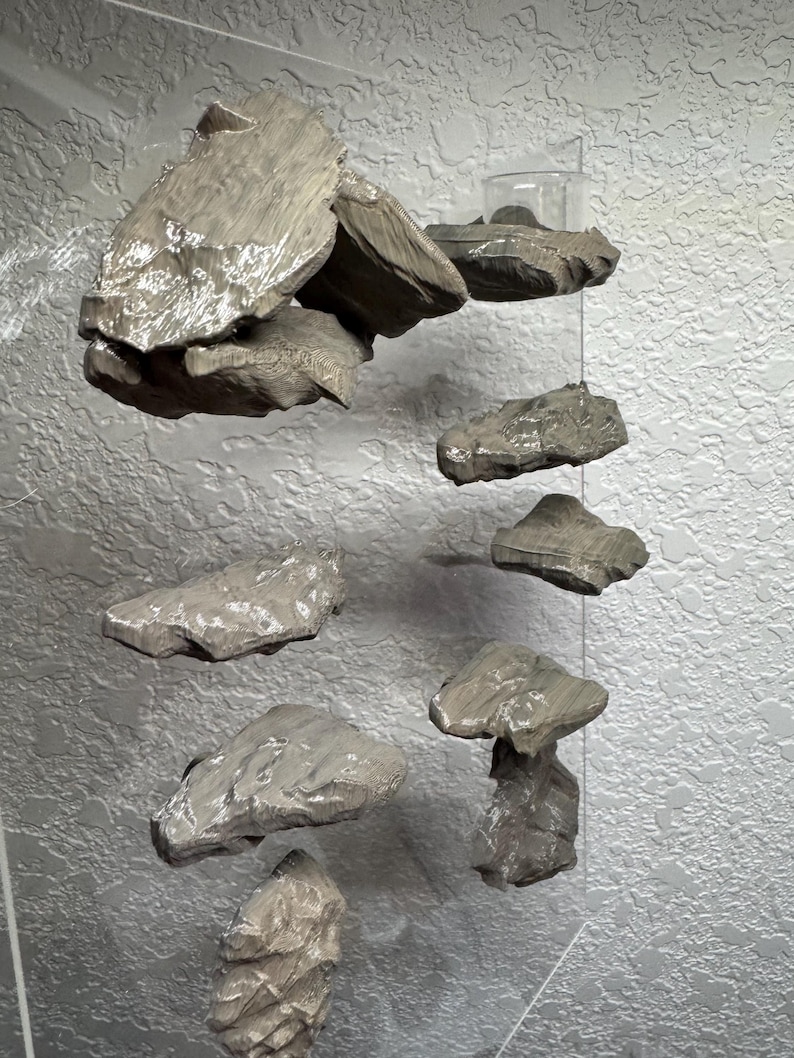 May include: A unique, modern wall art installation featuring several irregularly shaped, gray-toned stone pieces. The stones are suspended at varying heights, creating a floating effect against a textured, light gray background. The arrangement suggests a natural, organic theme.