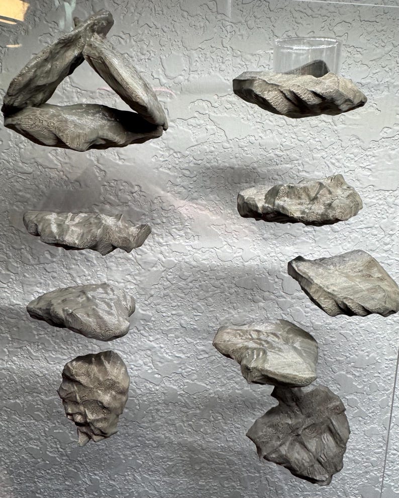 May include: A collection of gray, carved stone sculptures in various shapes and sizes. Some pieces are stacked, while others are displayed individually. The sculptures have a rough, textured surface and appear to be mounted on a textured wall.