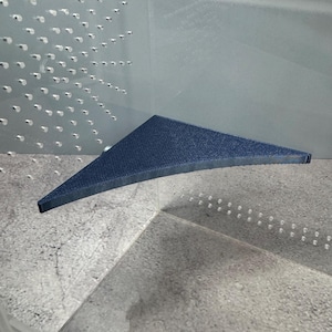 May include: A triangular, navy blue fabric object with a textured surface. The object is positioned in a clear, angular container with a gray, speckled base. The object's edges are dark blue.