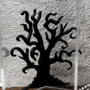May include: A black, stylized tree silhouette with twisted branches is displayed inside a clear, geometric box. The tree is set against a textured, off-white background. The overall design has a gothic or Halloween theme.