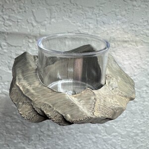 May include: A silver-colored votive candle holder with a clear glass cup. The holder has a textured, organic design. The candle holder is designed to hold a small tea light candle.