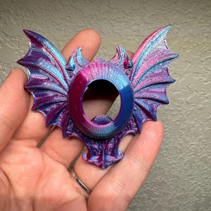 May include: A 3D printed decorative object featuring a central oval opening surrounded by intricate, wing-like structures. The piece displays a vibrant, iridescent color gradient shifting between shades of metallic blue, purple, and magenta.