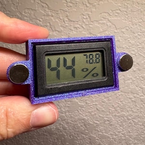 May include: A small, rectangular digital hygrometer with a purple frame and two silver magnetic attachments. The display shows the numbers "44" and "78.8%" in green. The device is held in a hand.