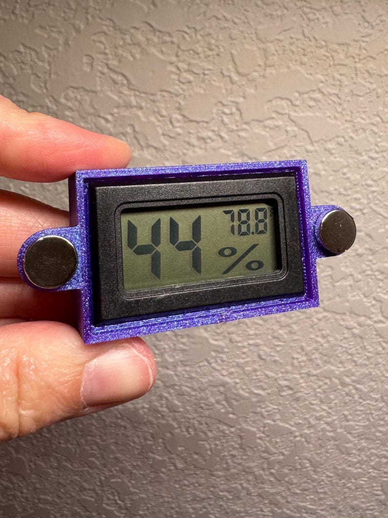 May include: A small, rectangular digital hygrometer with a purple frame and two silver magnetic attachments. The display shows the numbers "44" and "78.8%" in green. The device is held in a hand.
