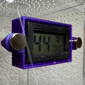 May include: A purple digital hygrometer with a black display showing 44% and 78.4%. The device has two silver magnetic attachments. The hygrometer is mounted inside a clear enclosure.